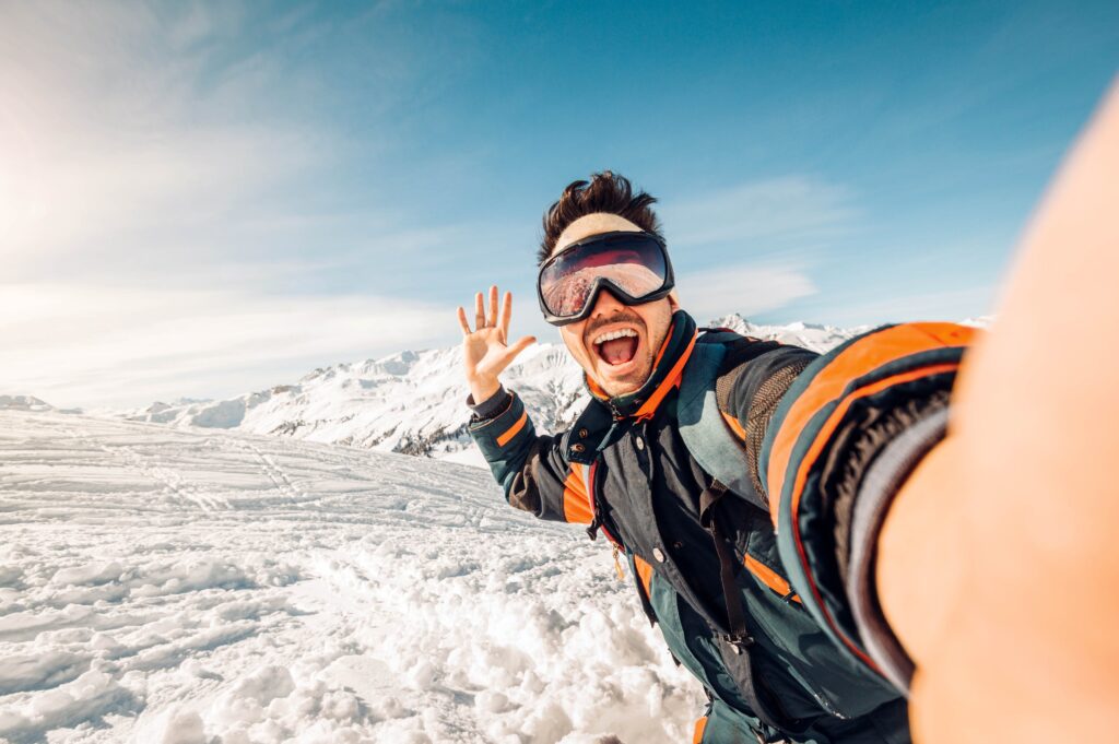 Eine Person mit Skibrille und Winterausrüstung macht ein Selfie auf einem verschneiten Berg, lächelt und winkt mit der Hand, während der klare blaue Himmel im Hintergrund ein beeindruckendes Gitter bildet.