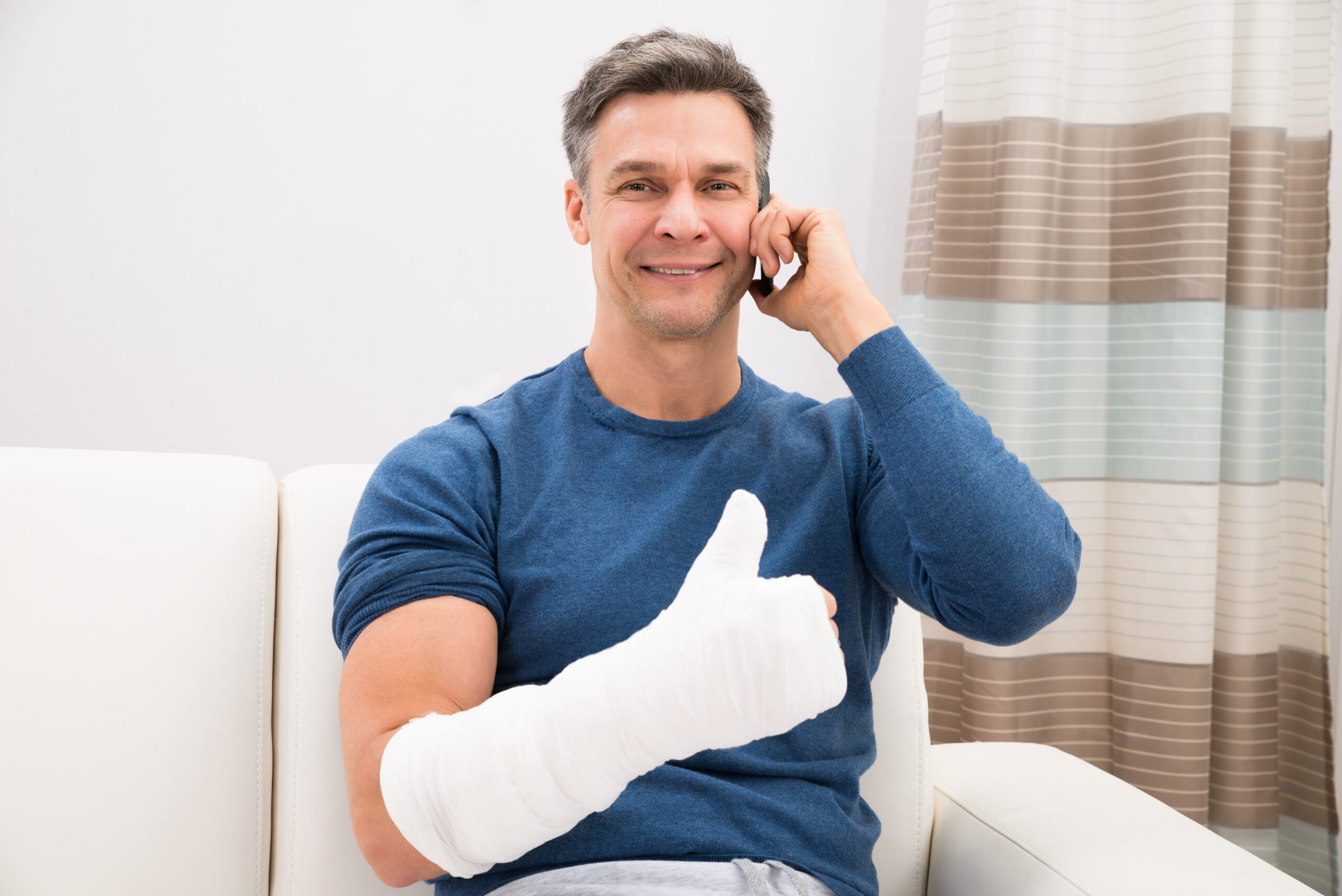 Ein Mann mit einem bandagierten Arm, wahrscheinlich von einer Schnittverletzung, sitzt auf einer Couch, zeigt den Daumen nach oben und telefoniert.
