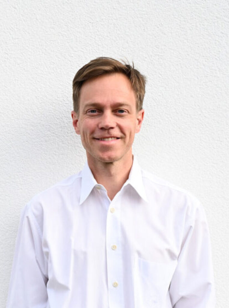 Ein Mann mit kurzen hellbraunen Haaren und weißem Hemd steht vor einer weißen, strukturierten Wand und lächelt in die Kamera - er repräsentiert die Handspezialisten Innsbruck.