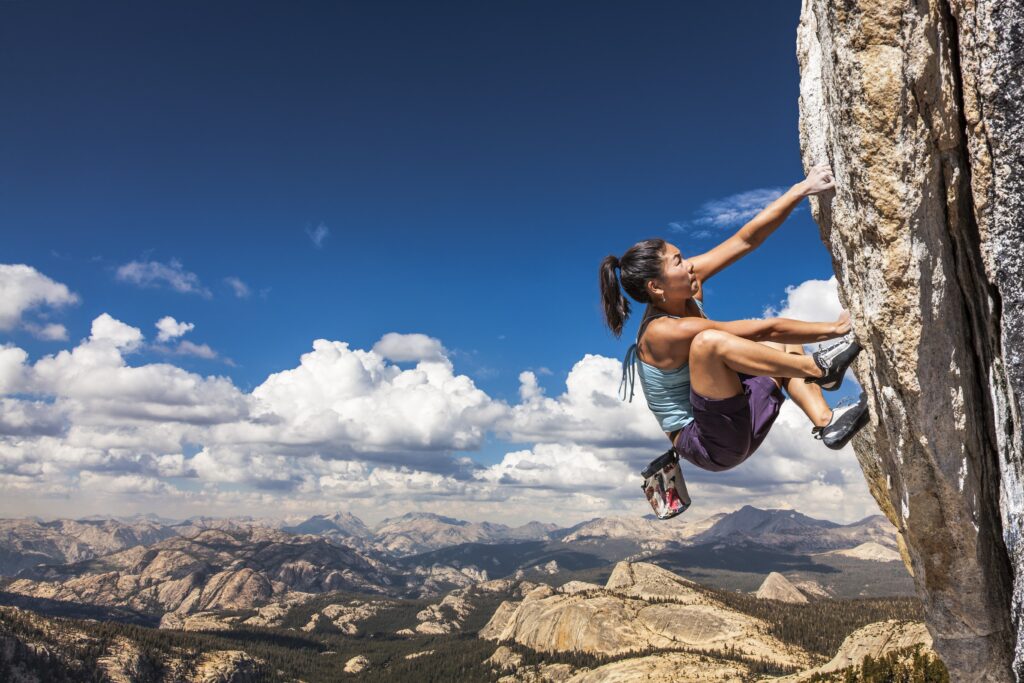 Eine Frau in Sportkleidung klettert eine steile Felswand hinauf, mit einer Berglandschaft und blauem Himmel im Hintergrund. Sie zeigt Geschicklichkeit und Entschlossenheit und hebt gleichzeitig die Bedeutung der Vorbeugung von Kletterverletzungen wie einem A2-Ringbandriss hervor.
