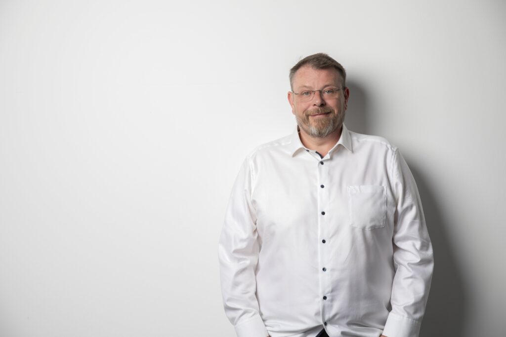 Vor einer schlichten weißen Wand steht ein Mann mit Brille und Bart, der ein weißes Button-up-Hemd mit dunklen Knöpfen trägt - das sympathische Erscheinungsbild des Teams der Handspezialisten Innsbruck.
