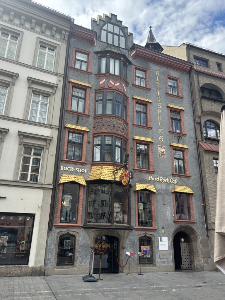 Vierstöckiges Gebäude mit dekorativen roten und goldenen Akzenten, das im Erdgeschoss ein Hard Rock Cafe und einen Rock Shop beherbergt und sich in einer Fußgängerzone in der Nähe der Handspezialisten Innsbruck befindet.