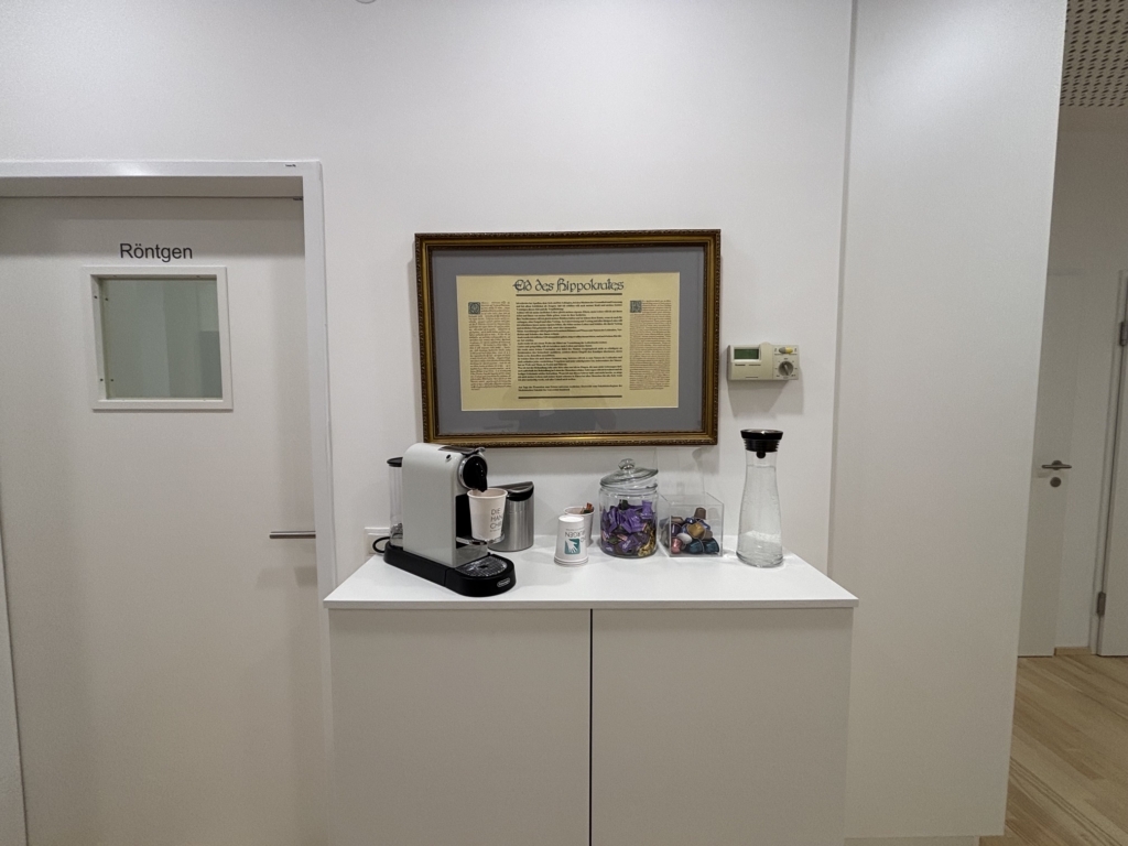 Eine kleine Kaffeestation mit Kaffeemaschine, Tassen, Zucker, Wasserkaraffe und einer Dose mit Kaffeepads steht an einer weißen Wand unter einem gerahmten Dokument bei den Handspezialisten Innsbruck. Auf der linken Seite befindet sich eine Tür mit der Aufschrift "Röntgen".
