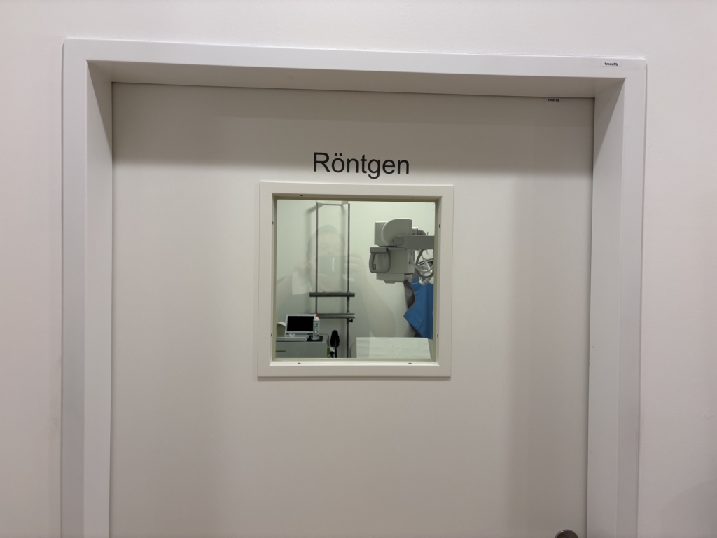 Eine geschlossene Tür mit der Aufschrift "Röntgen" und einem kleinen Fenster, das den Blick auf medizinische Geräte und eine Person in Schutzkleidung freigibt, markiert den Fachbereich der Handspezialisten Innsbruck.