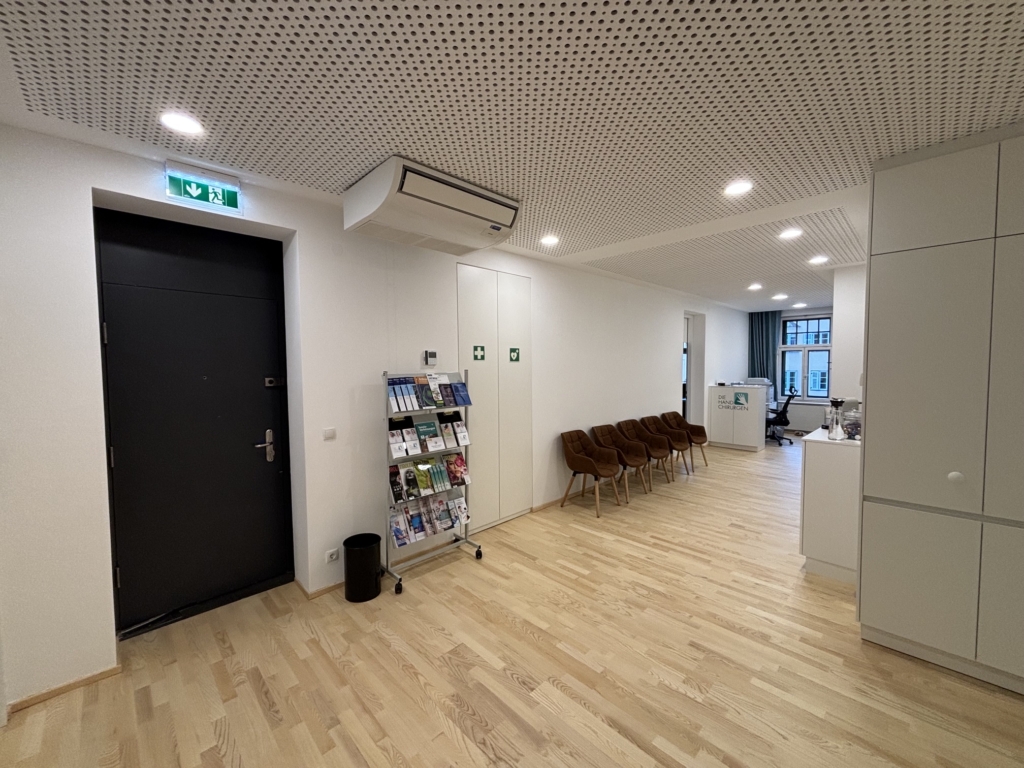 Ein moderner Büroflur mit Holzböden, braunen Stühlen an der Wand, einem Prospektständer neben einer schwarzen Tür und sichtbaren Notausgangsschildern - ein einladender Raum für die Besucher der Handspezialisten Innsbruck.