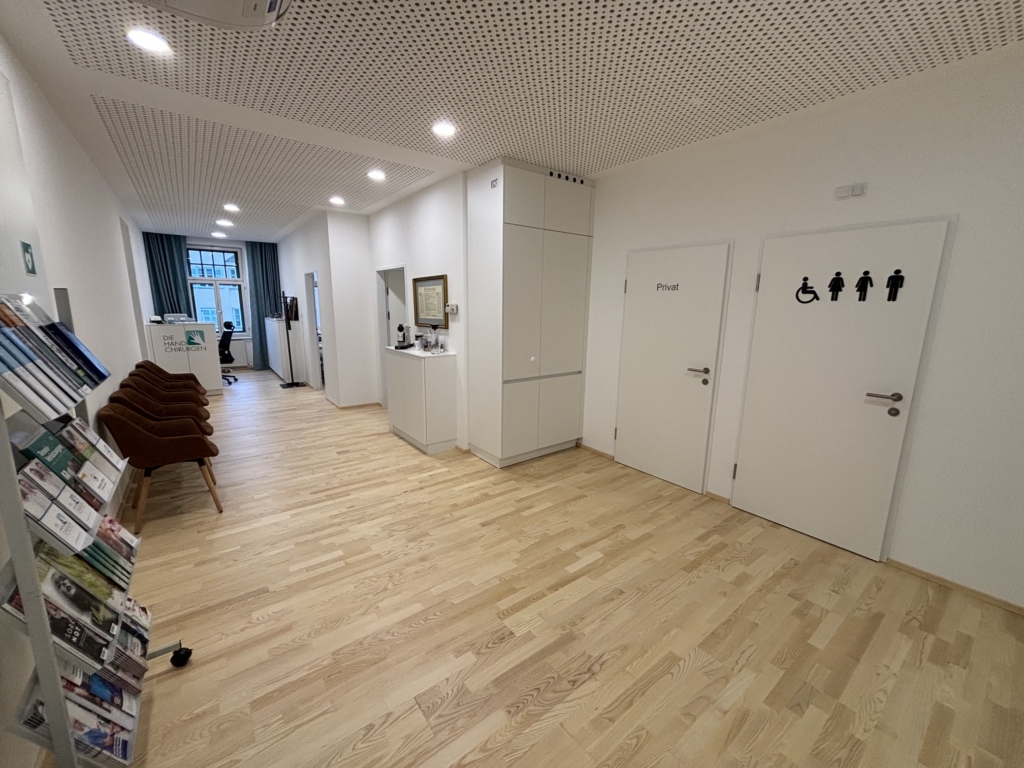 Ein sauberer, moderner Klinikflur bei den Handspezialisten Innsbruck ist mit Holzböden, braunen Stühlen, einem Zeitschriftenständer und zwei Toilettentüren ausgestattet - eine mit der Aufschrift "Privat" und die andere mit barrierefreien und geschlechtsneutralen Symbolen.