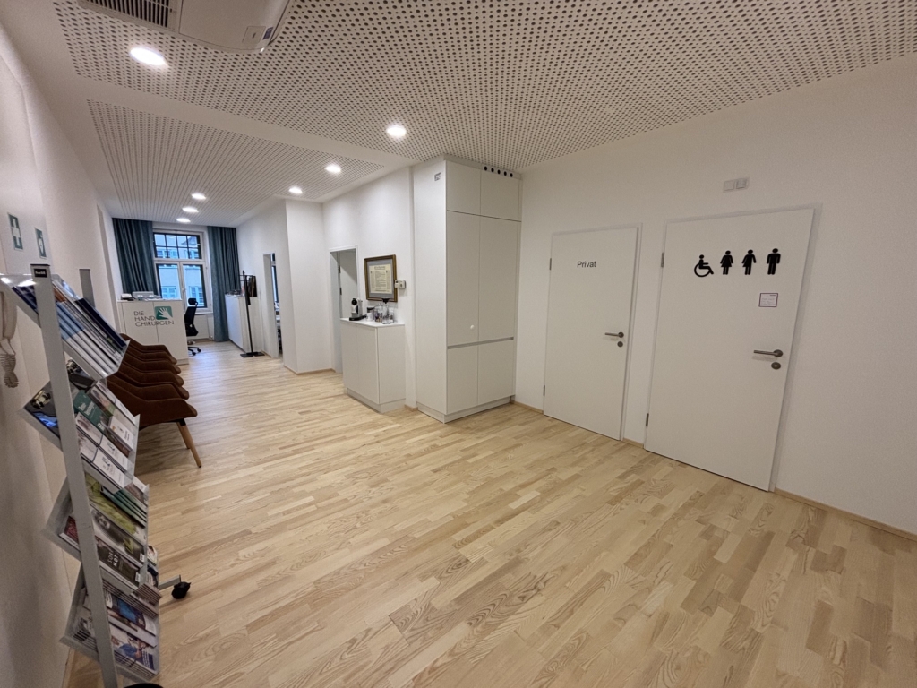 Ein sauberer, moderner Flur bei den Handspezialisten Innsbruck mit Holzböden, Informationsbroschüren auf einem Regal, braunen Stühlen und Türen, die mit Toiletten und einem privaten Raum beschriftet sind. Im Hintergrund sitzt eine Person an einem Schreibtisch.